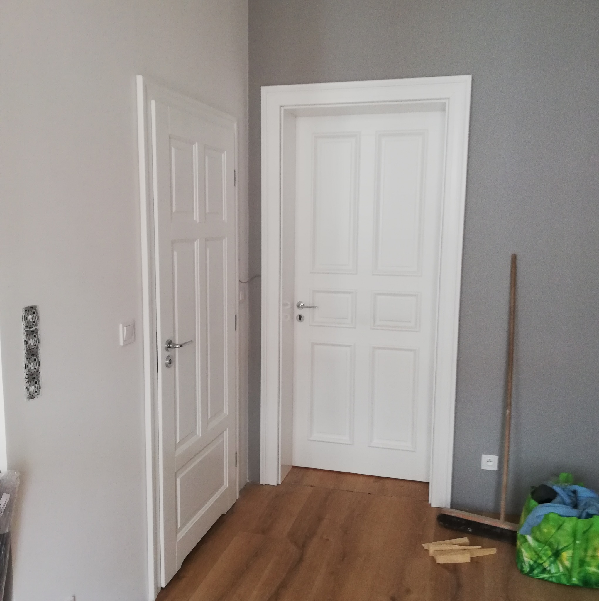 Dwa białe, panelowe skrzydła drzwiowe, jedno otwarte, drugie zamknięte, na drewnianej podłodze. Szara ściana w tle. Widoczne gniazdka elektryczne i listwy przypodłogowe.