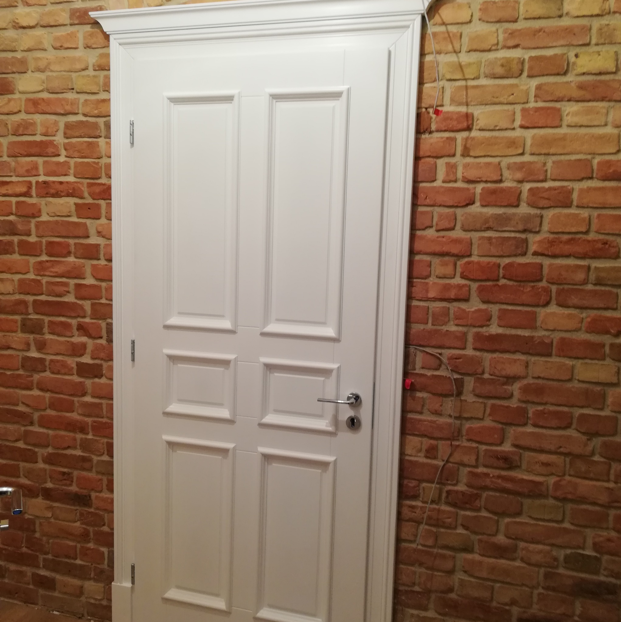 Klasyczne białe drzwi wewnętrzne z dekoracyjną ramą, zamontowane w ścianie z czerwonej cegły. Widoczne zawiasy i klamka.