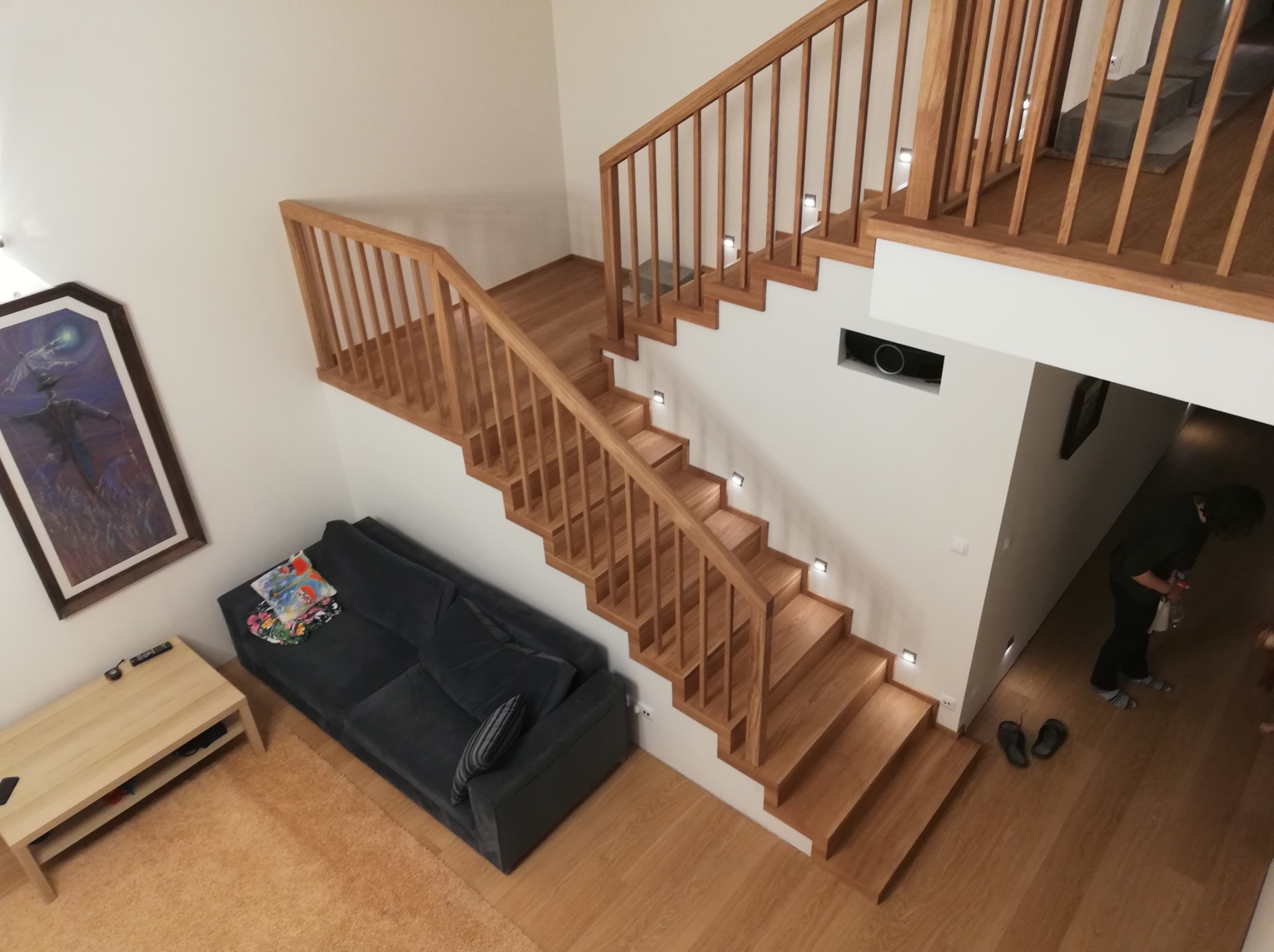 Jasne, drewniane schody z balustradą i oświetleniem LED, widziane z góry. Obok schodów szara sofa i obraz na ścianie. Parkiet na podłodze.