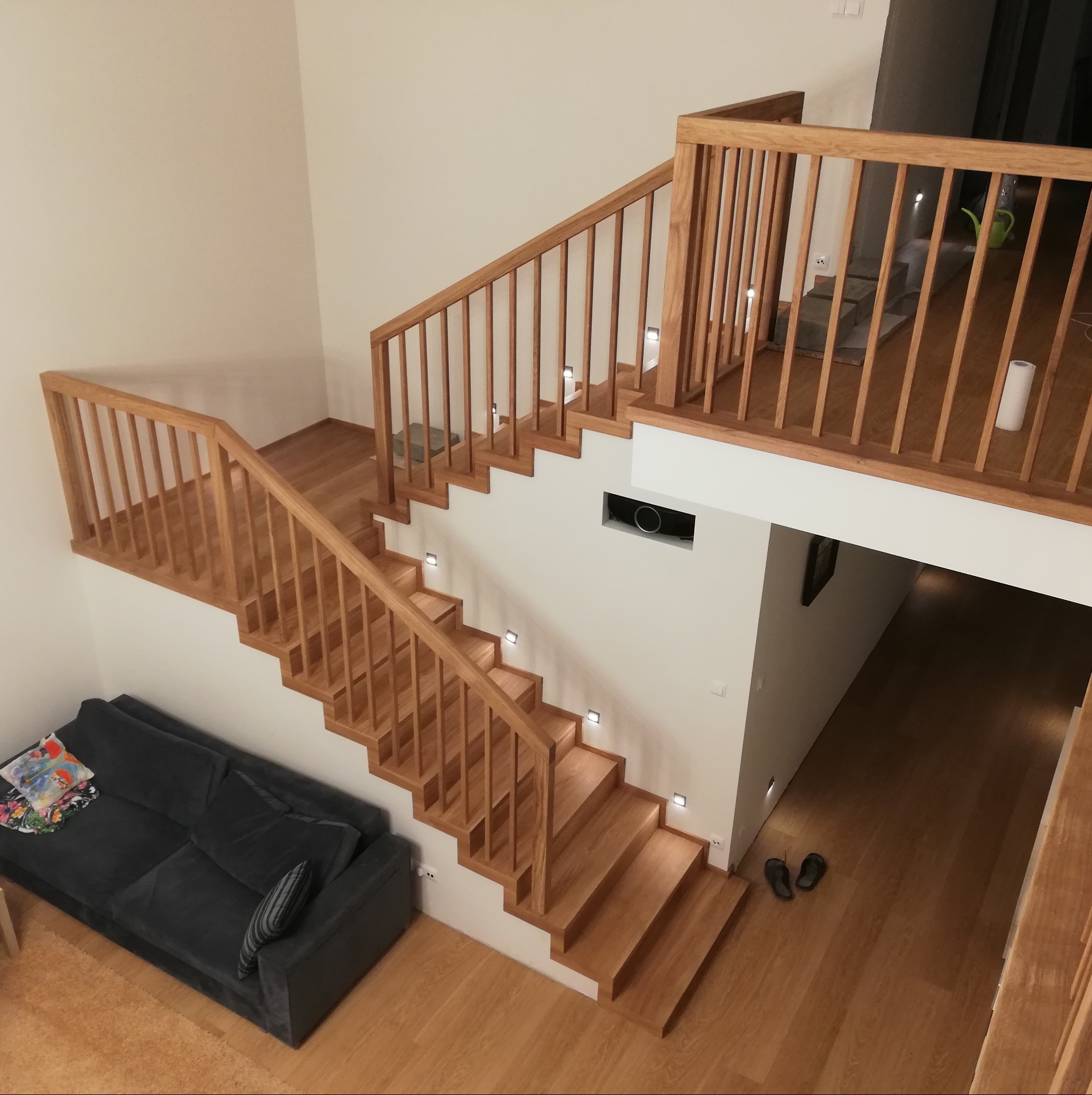 Dwupoziomowe, jasne schody z drewnianą balustradą, oświetlone punktowymi lampami, widziane z góry, z szarą sofą i drewnianą podłogą w tle.