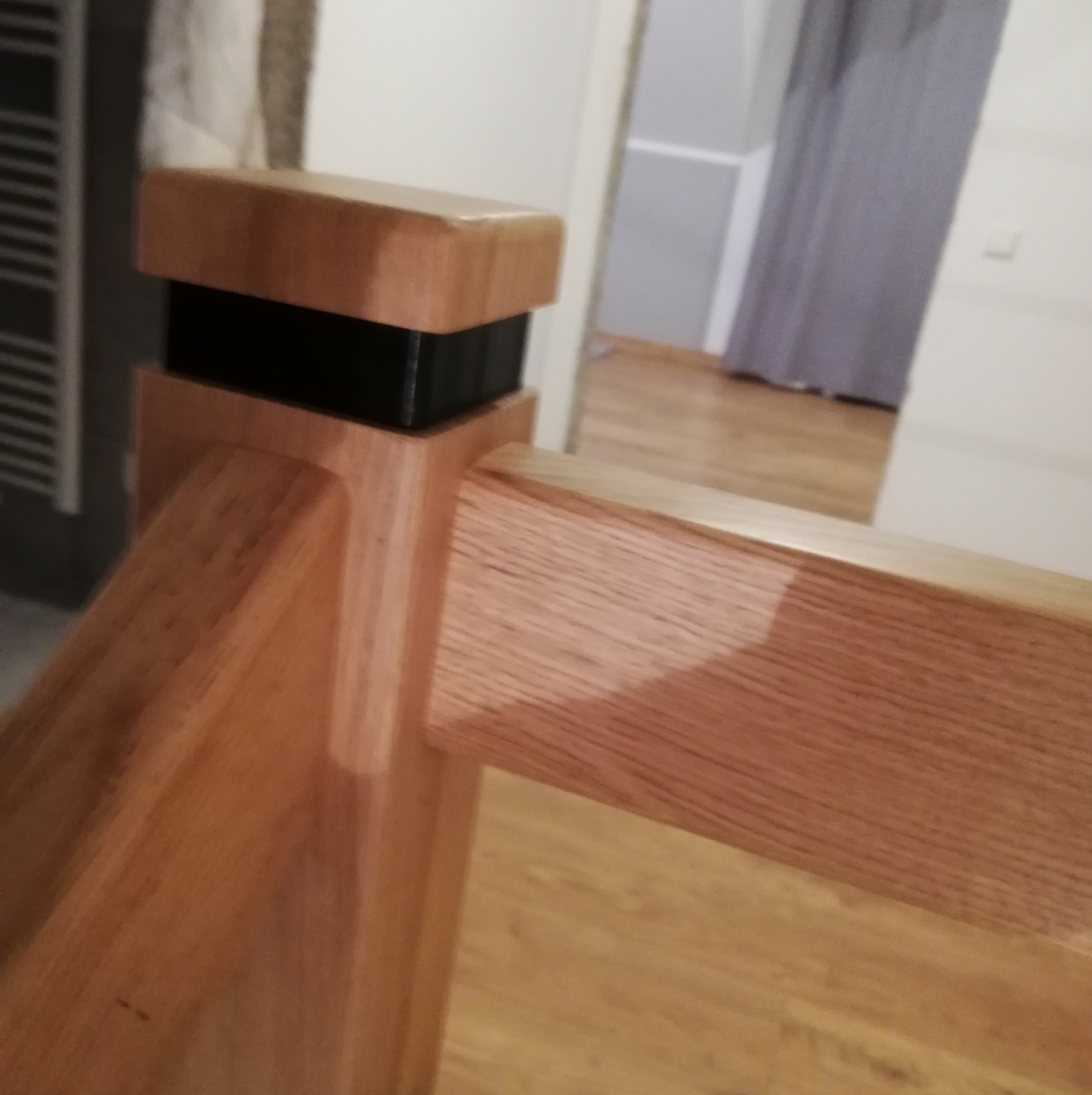 Fragment drewnianej balustrady z czarnym łącznikiem, widoczny parkiet i fragment zasłony w tle. Ujęcie z bliska podkreśla detale wykonania.