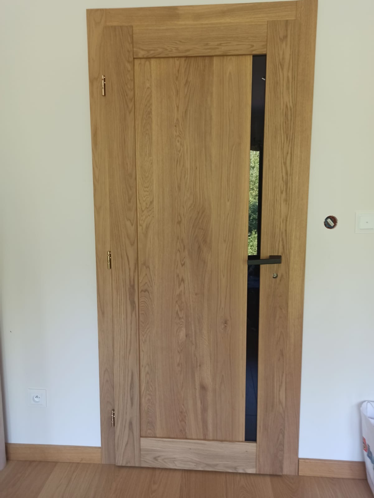 Jasne, dębowe drzwi wewnętrzne z czarnym, wąskim przeszkleniem, zamontowane w Starachowicach. Widoczne zawiasy i klamka, podłoga drewniana.