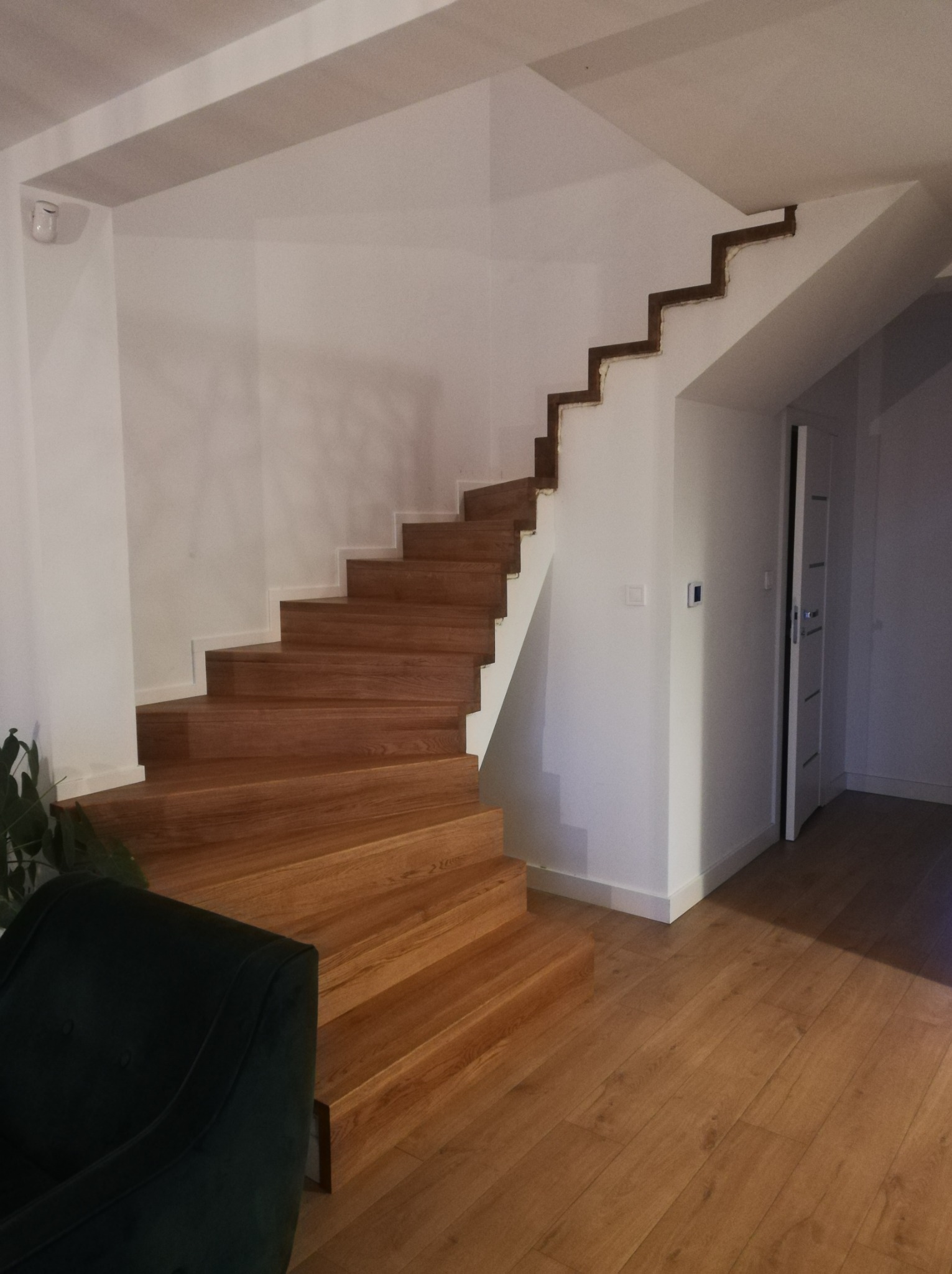 Jasne, drewniane schody w nowoczesnym wnętrzu z białymi ścianami i podłogą. Widoczny fragment zielonego fotela w lewym dolnym rogu kadru.