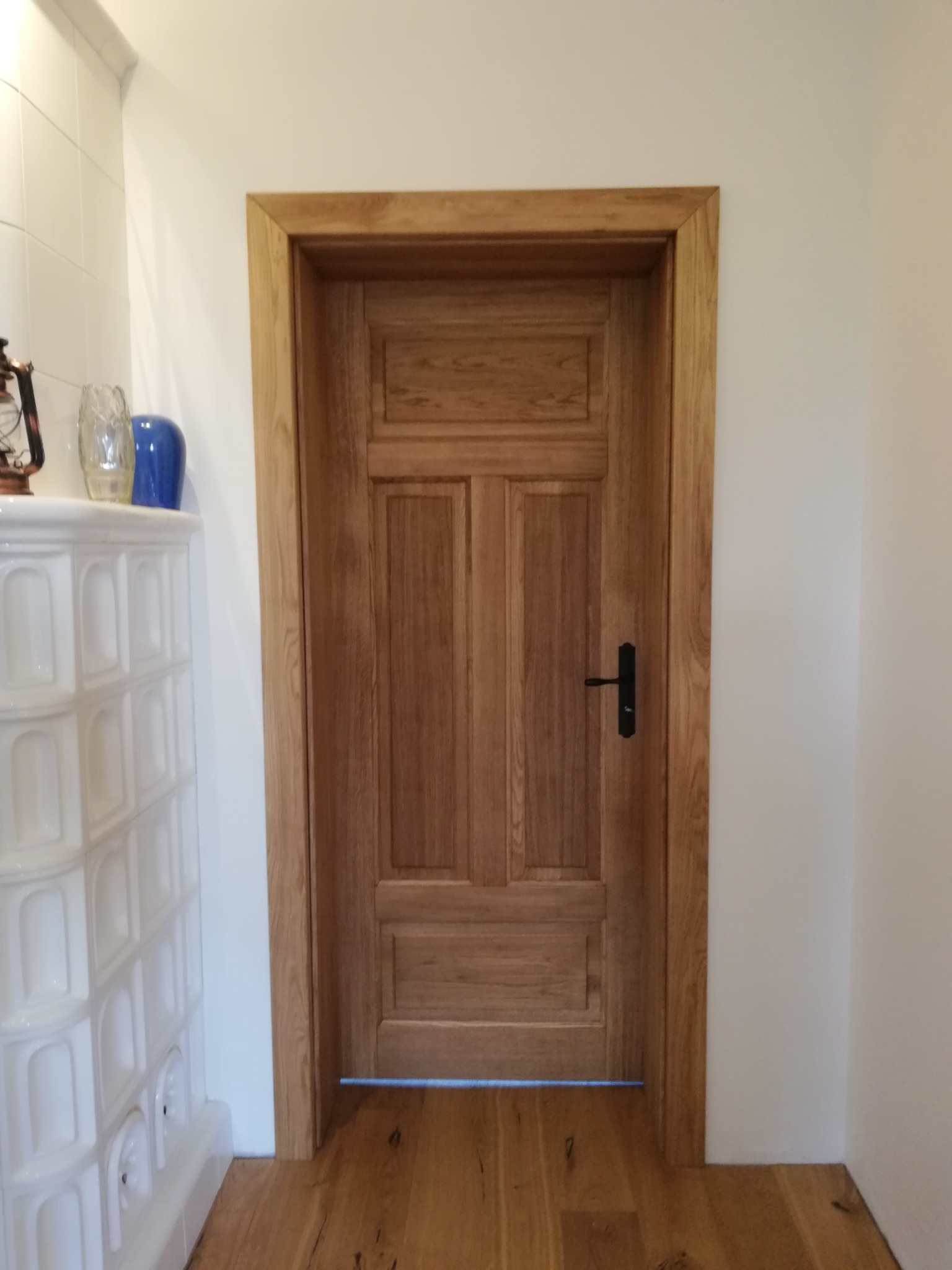 Klasyczne, drewniane drzwi wewnętrzne z czarną klamką, wkomponowane w białe ściany, z widoczną podłogą z desek i ozdobnym piecem kaflowym w tle.