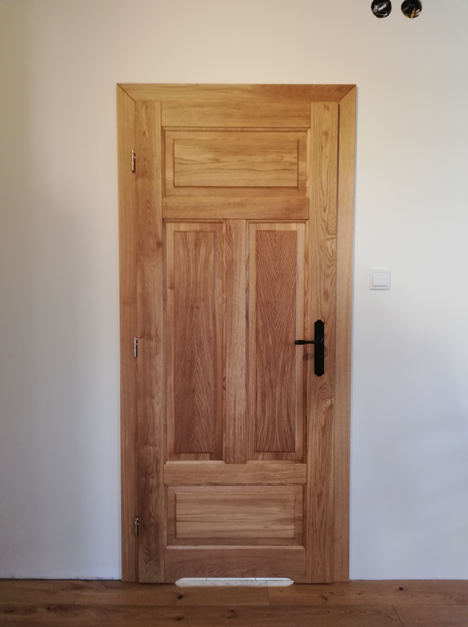 Dębowe drzwi wewnętrzne z czarną klamką, widoczne zawiasy i fragment drewnianej podłogi. Świeżo zamontowane, na ścianie widoczne puszki elektryczne i włącznik światła.