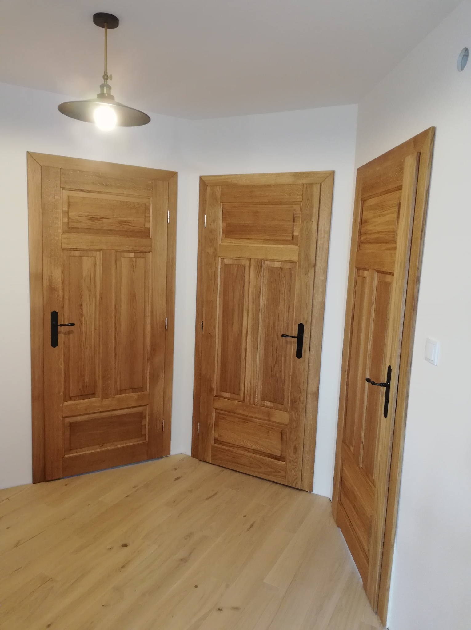 Trzy drewniane drzwi wewnętrzne z czarnymi klamkami, oświetlone lampą wiszącą, ukazane z perspektywy podłogi, eksponują naturalną fakturę drewna.