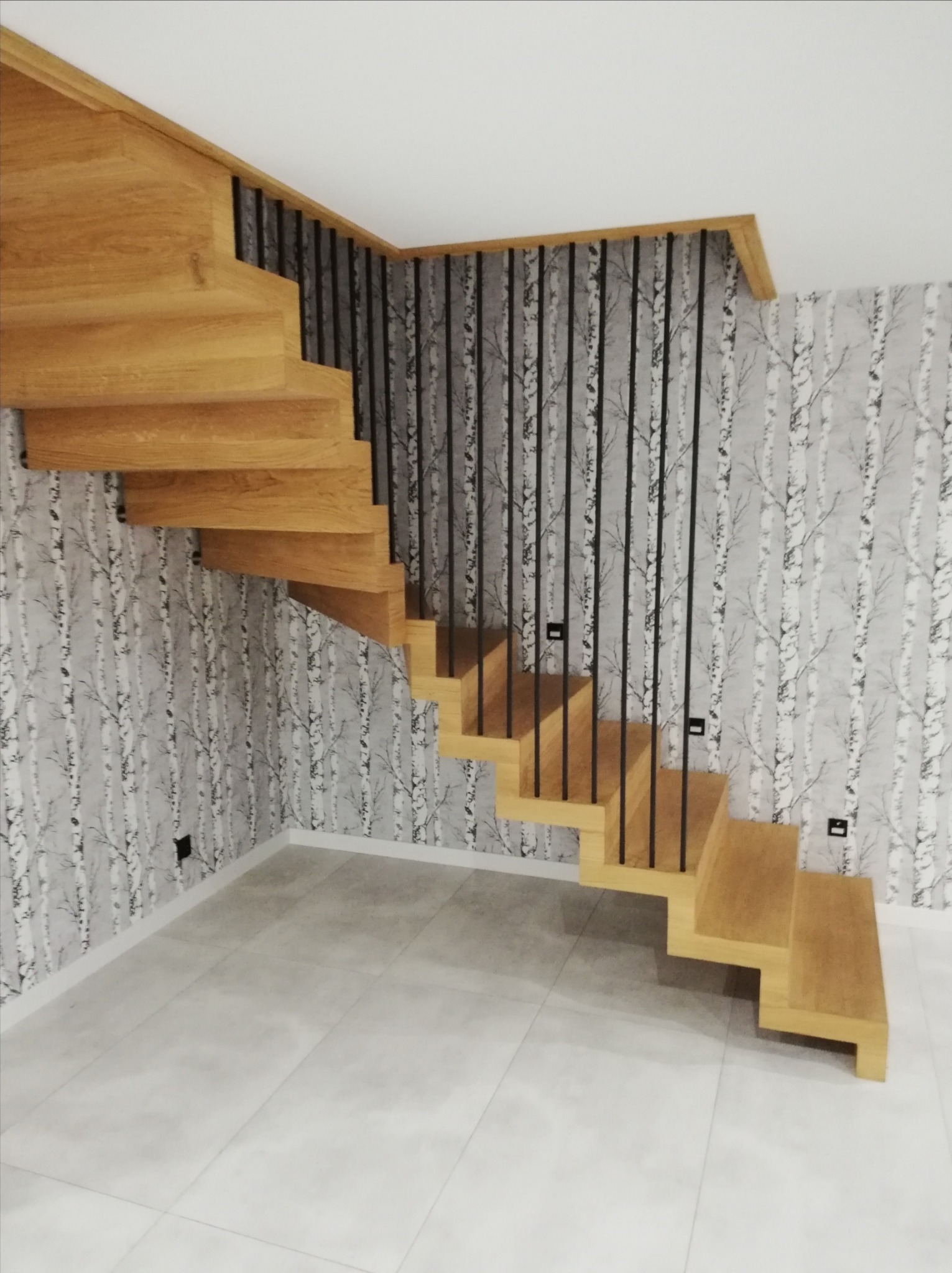 Drewniane schody z czarną balustradą na tle tapety w brzozy. Stopnie z litego drewna, nowoczesny design, jasne płytki podłogowe. Widok z dołu na bieg schodów.