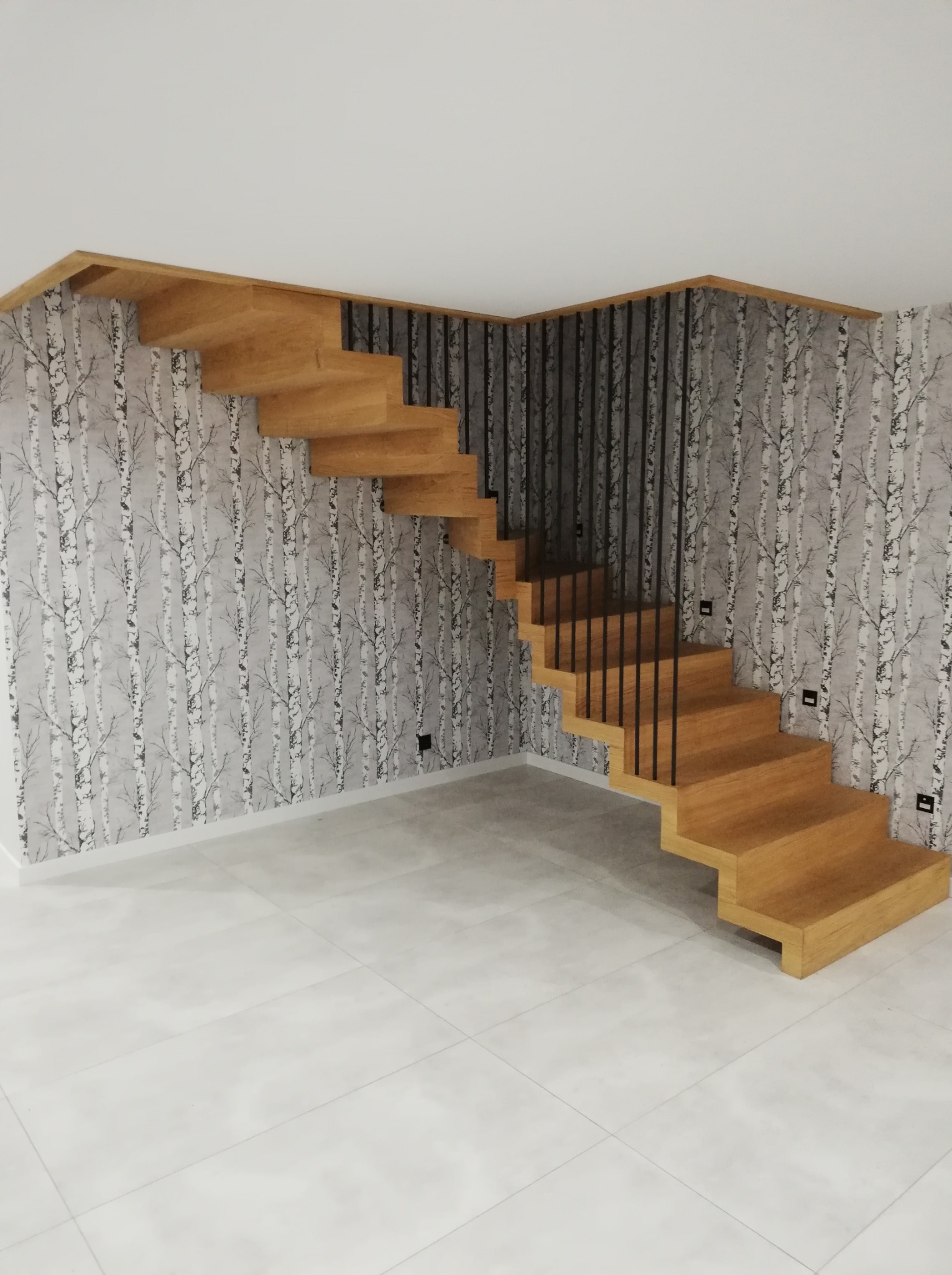 Nowoczesne, ażurowe schody z balustradą w Starachowicach. Stopnie z jasnego drewna kontrastują z ciemnymi, pionowymi elementami balustrady i tapetą w motywie brzozy.