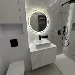Mateusz sadecki - Nowoczesna łazienka z okrągłym lustrem LED, białą umywalką na szafce z frezowanymi frontami i kabiną prysznicową typu walk-in. Marmurowe płytki i czarne akcenty.