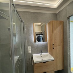 Mateusz sadecki - Nowoczesna łazienka z szarą glazurą, kabiną prysznicową ze szkła, szafką z umywalką i lustrem z podświetleniem LED. W lustrze odbija się pralka.