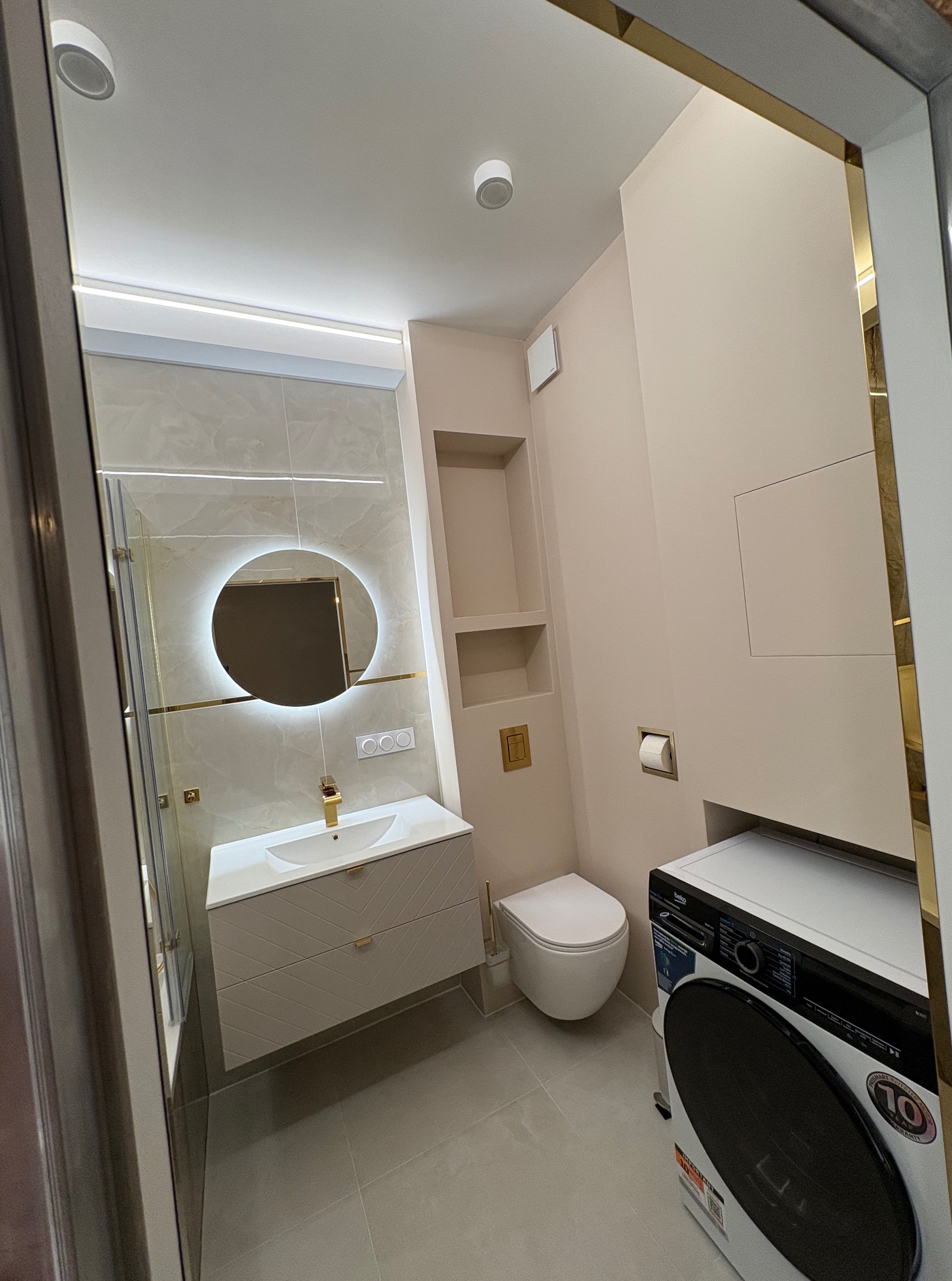Nowoczesna łazienka w beżach: okrągłe lustro z podświetleniem, biała umywalka z szafką w geometryczne wzory, toaleta i pralka. Złote akcenty w wykończeniu.