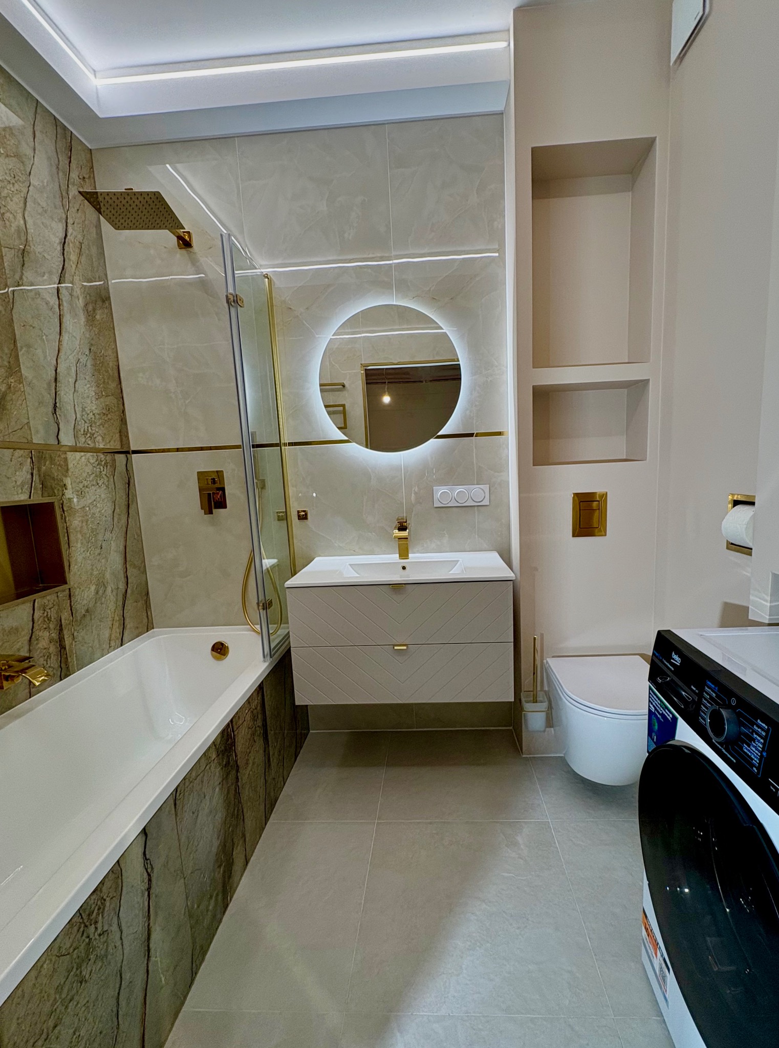 Wykończona łazienka w beżach z akcentami złota: kabina walk-in, umywalka z szafką w jodełkę, podświetlane lustro i pralka. Nowoczesny design.