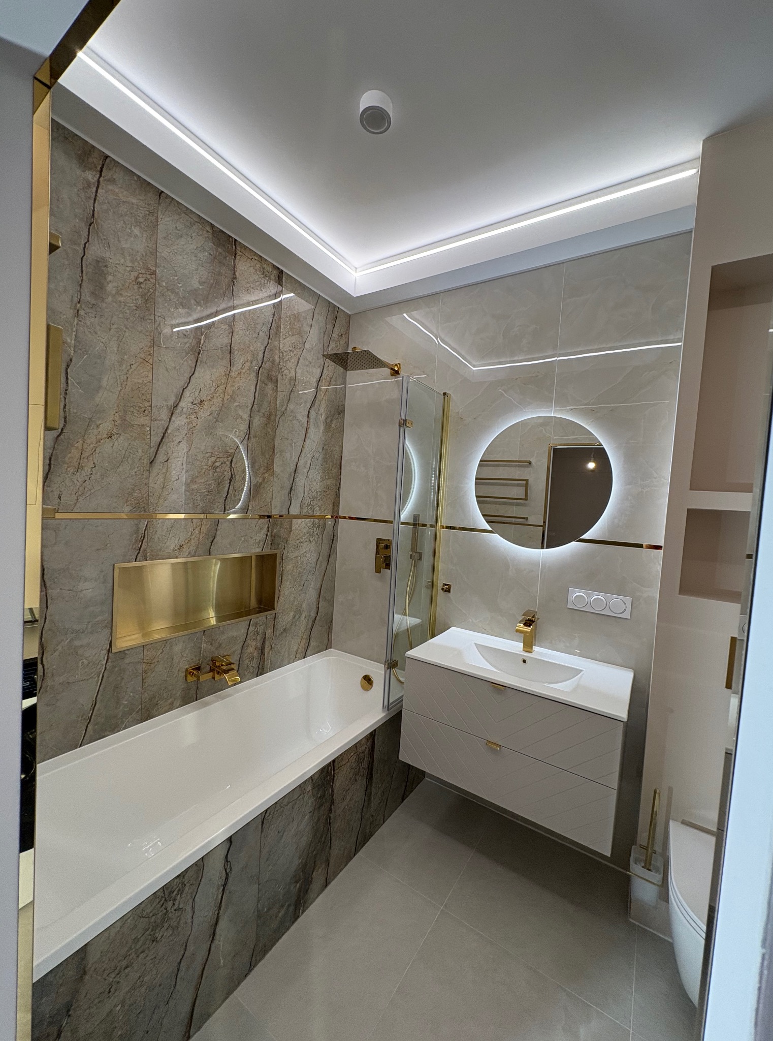 Elegancka łazienka z marmurowymi płytkami, złotymi akcentami i podświetlanym lustrem. Nowoczesna armatura i oświetlenie LED tworzą luksusowy klimat.