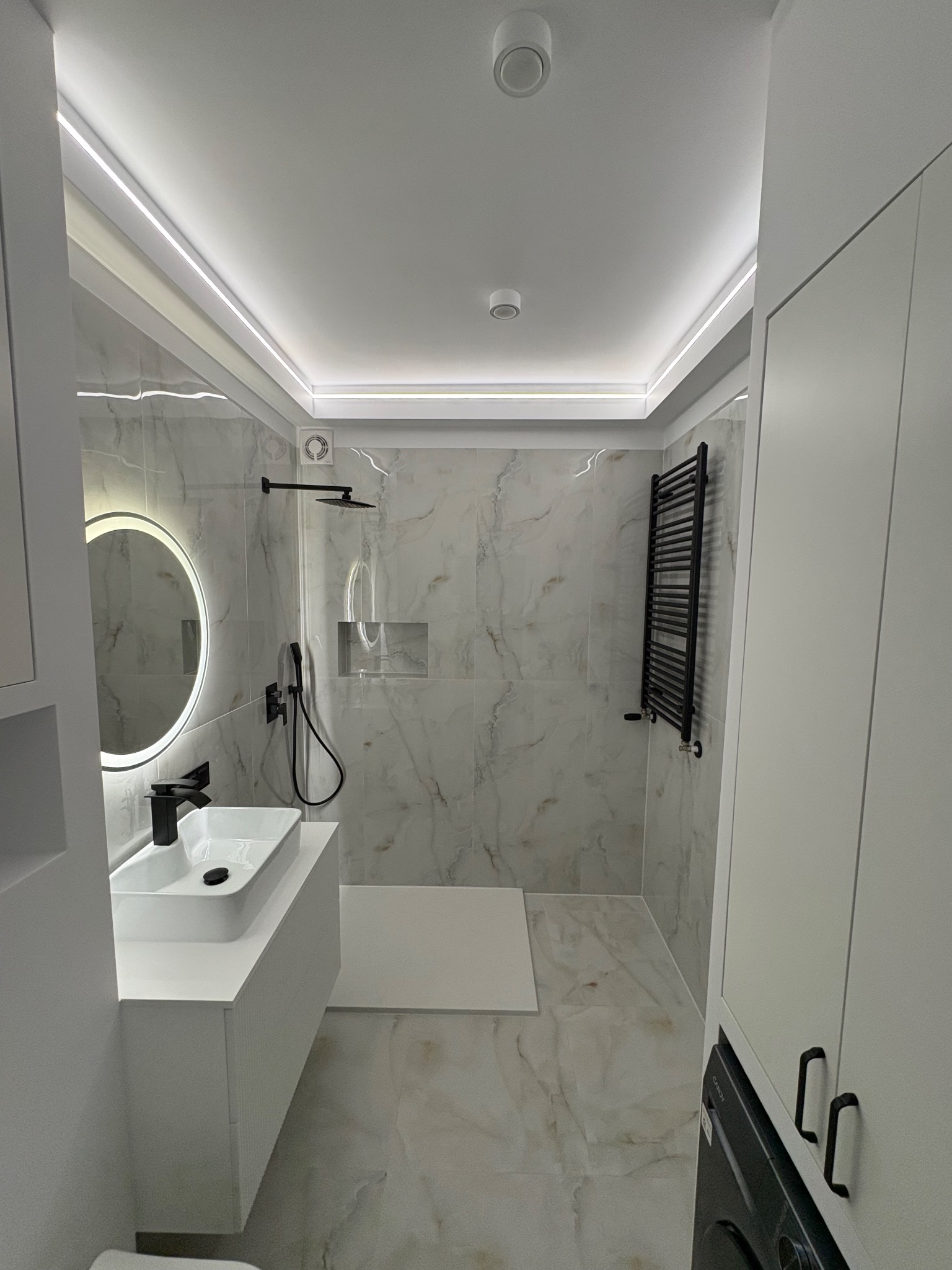 Nowoczesna łazienka z marmurowymi płytkami, czarną armaturą i podświetlanym lustrem. Minimalistyczny design z akcentami czerni i bieli, oświetlenie LED.