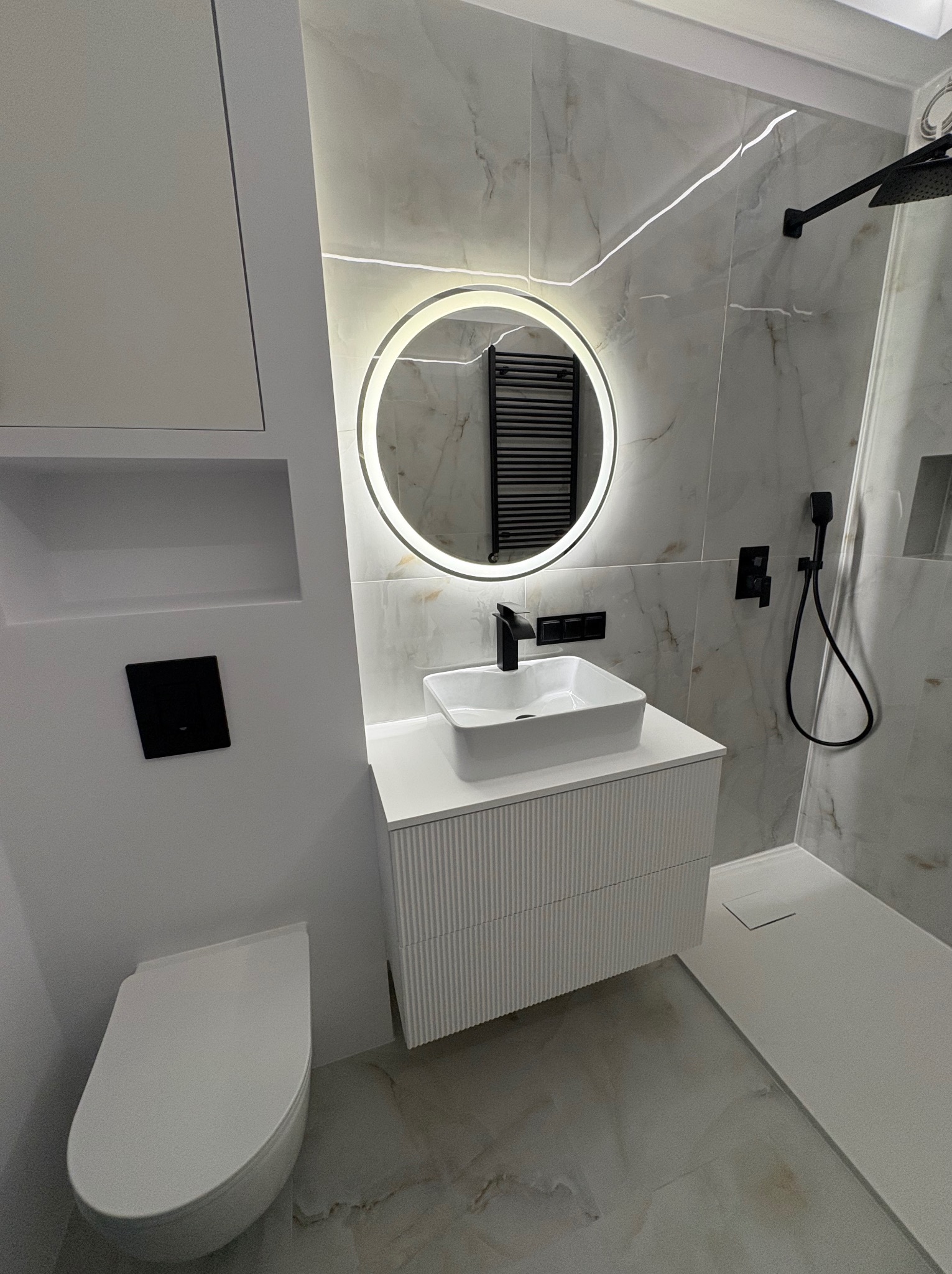 Nowoczesna łazienka z okrągłym lustrem LED, białą umywalką na szafce z frezowanymi frontami i kabiną prysznicową typu walk-in. Marmurowe płytki i czarne akcenty.
