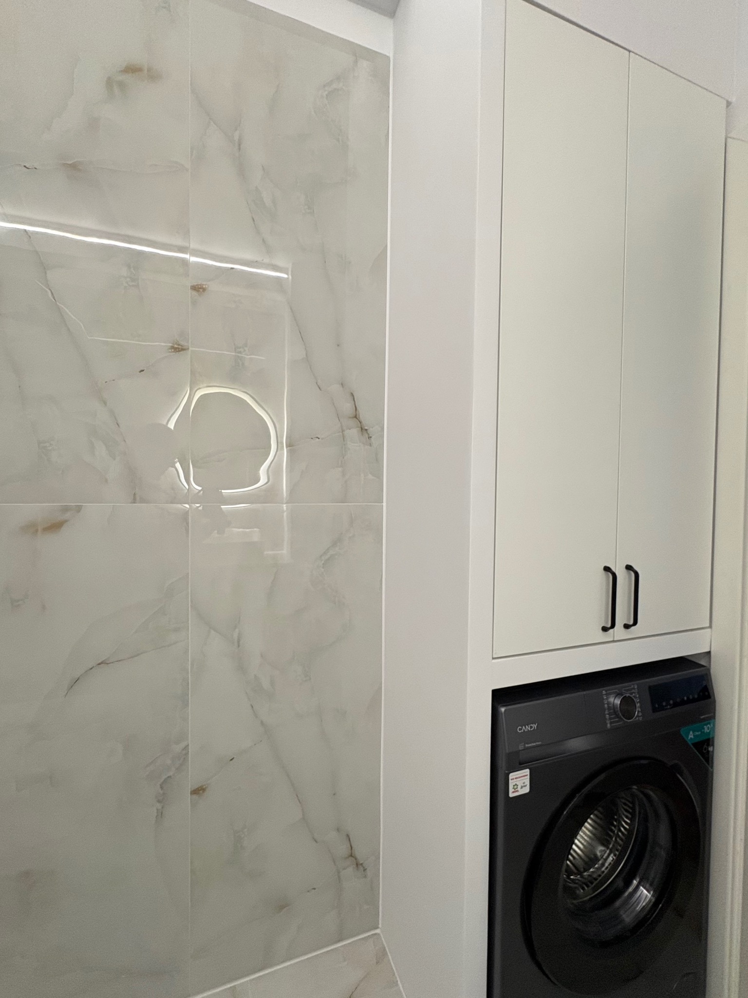 Biała szafka łazienkowa nad pralką, marmurowe płytki na ścianie z subtelnym oświetleniem LED. Minimalistyczny design, czarne uchwyty szafek.