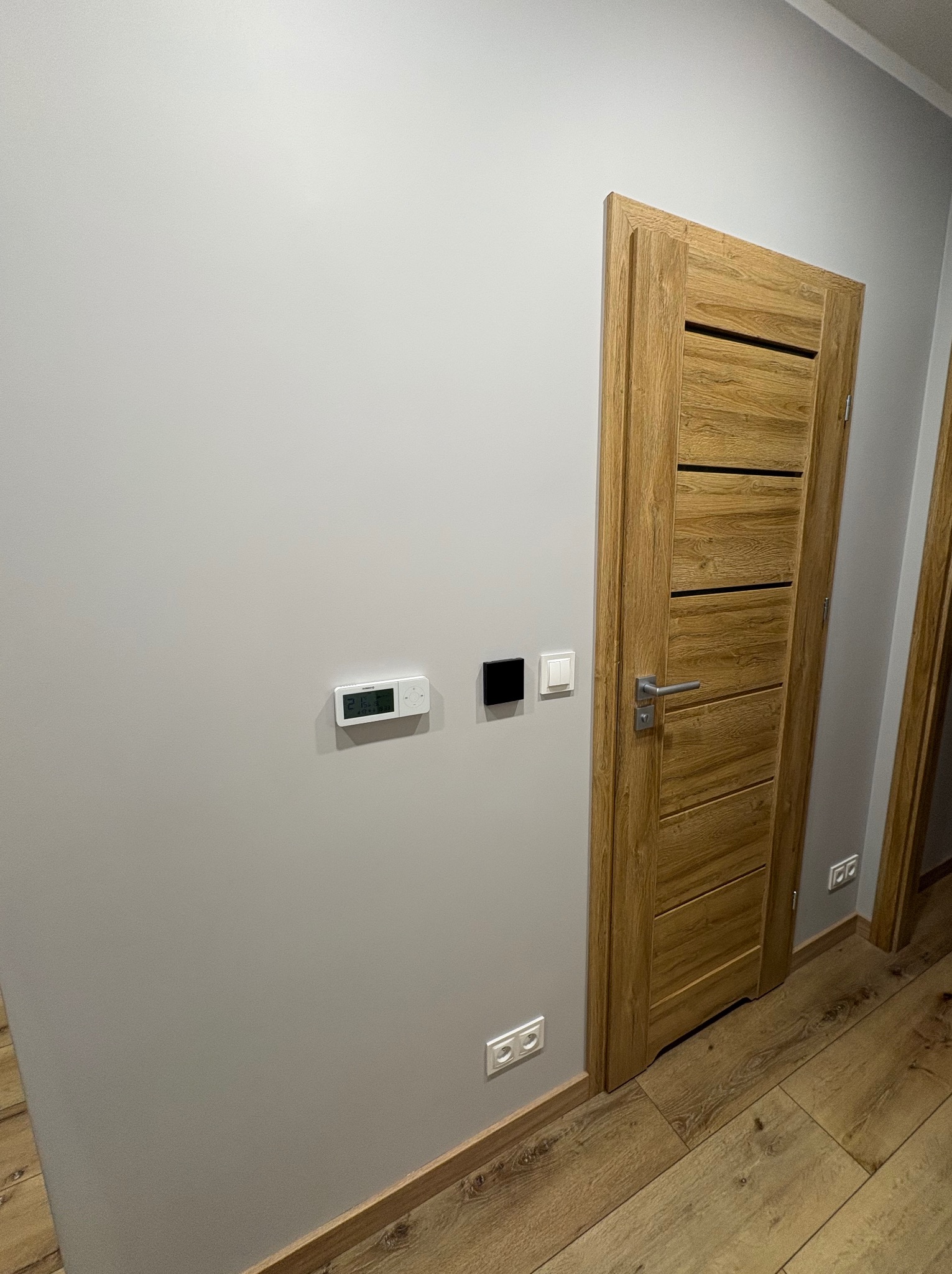 Zamknięte, drewniane drzwi wewnętrzne z poziomymi wstawkami, zamontowane w szarej ścianie. Widoczny termostat, włącznik światła i gniazdko elektryczne. Podłoga drewniana.