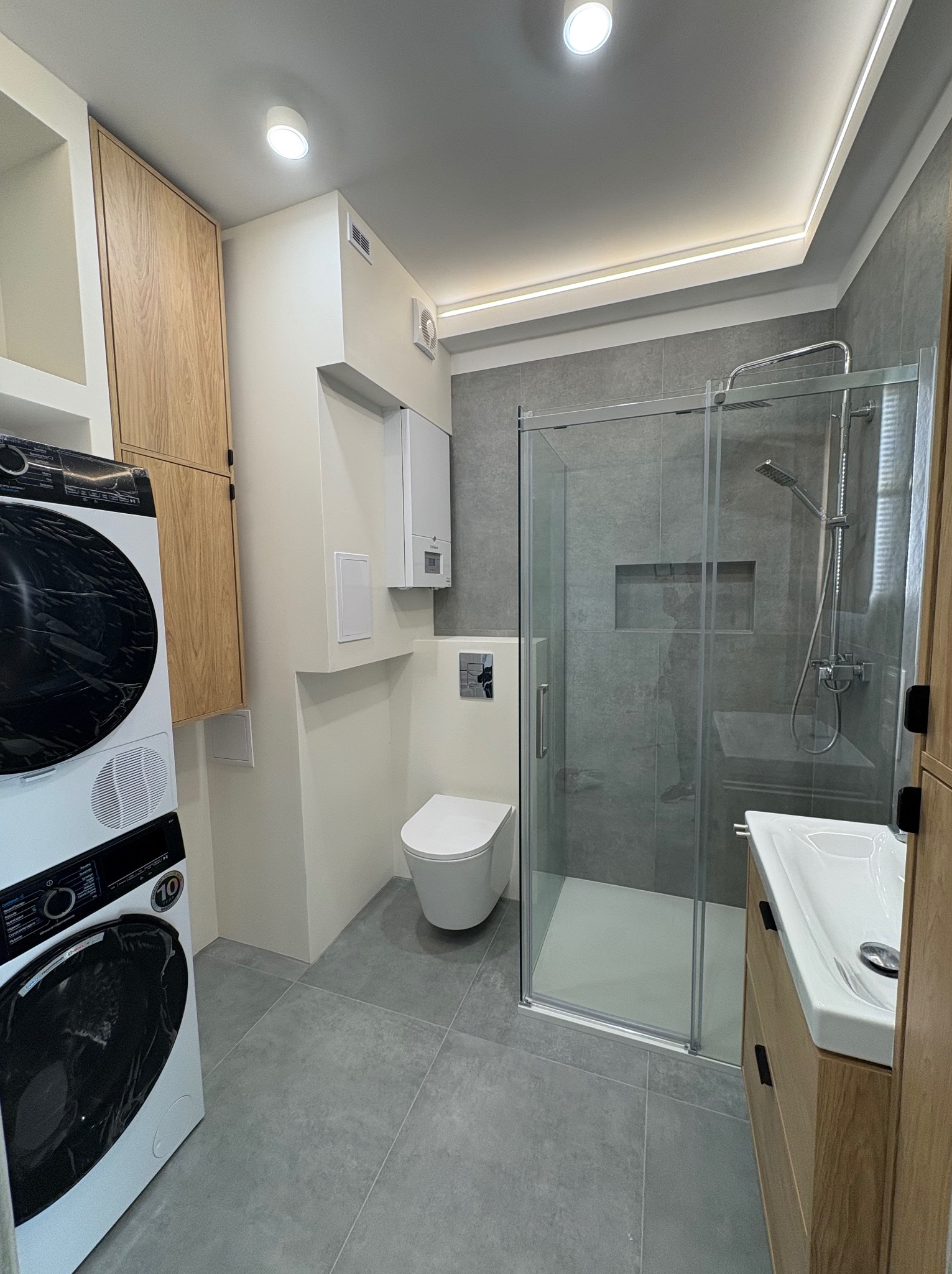 Nowoczesna łazienka z szarą glazurą, kabiną prysznicową typu walk-in, toaletą podwieszaną i pralko-suszarką z czarnym frontem. Oświetlenie LED podkreśla minimalistyczny design.