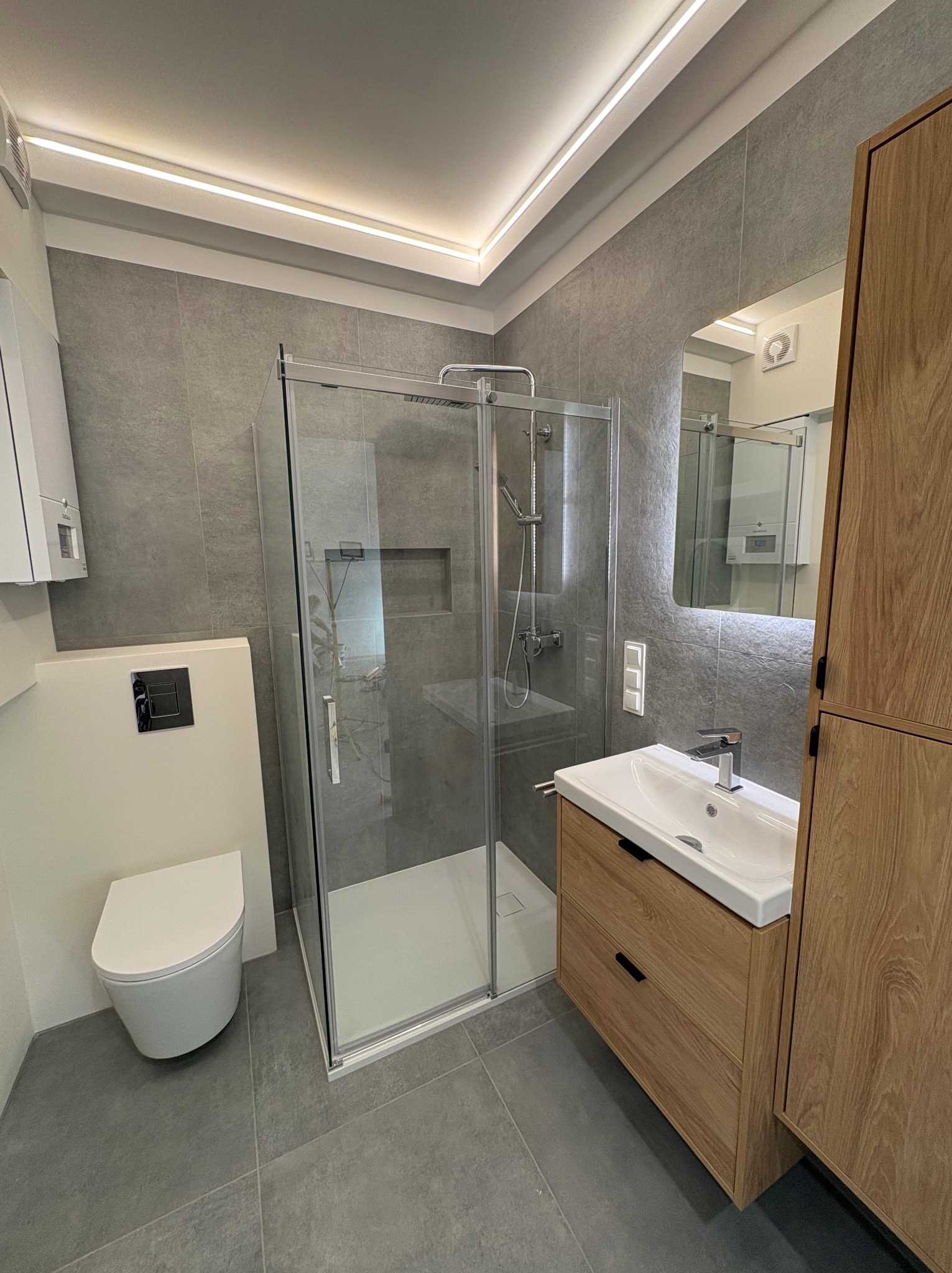Nowoczesna łazienka z kabiną prysznicową, umywalką z szafką z jasnego drewna i podwieszaną toaletą. Szare płytki na ścianach i podłodze, oświetlenie LED.