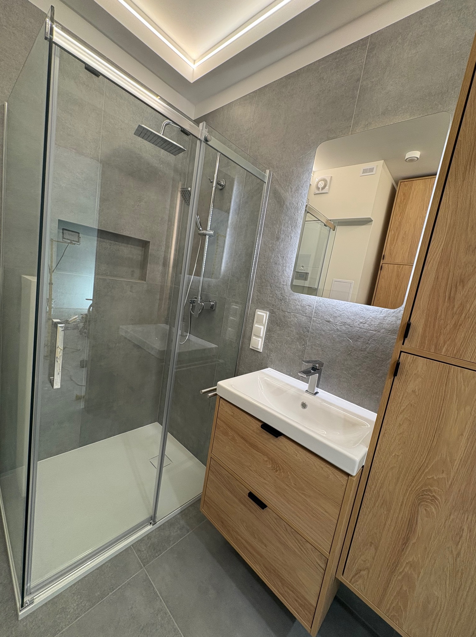 Nowoczesna łazienka z kabiną prysznicową typu walk-in, umywalką na szafce z drewnianym fornirem i dużym lustrem. Szare płytki na ścianach i podłodze, oświetlenie LED.
