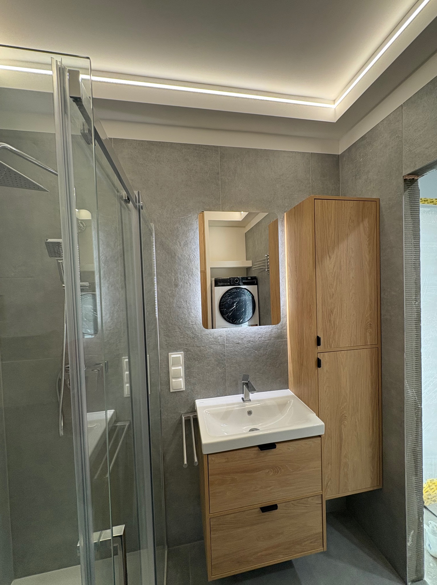 Nowoczesna łazienka z szarą glazurą, kabiną prysznicową ze szkła, szafką z umywalką i lustrem z podświetleniem LED. W lustrze odbija się pralka.