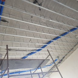 Jakub Kreft - Montaż sufitu podwieszanego z widocznymi elementami konstrukcyjnymi i niebieskimi rurami instalacyjnymi. Widok z dołu, rusztowanie w kadrze.