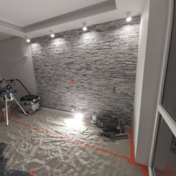 Jakub Kreft - Wykończona ściana z kamienia dekoracyjnego w pokoju, zabezpieczona folią podłoga, narzędzia i sprzęt do cięcia płytek widoczne w tle. Wnętrze w trakcie prac wykończeniowych.