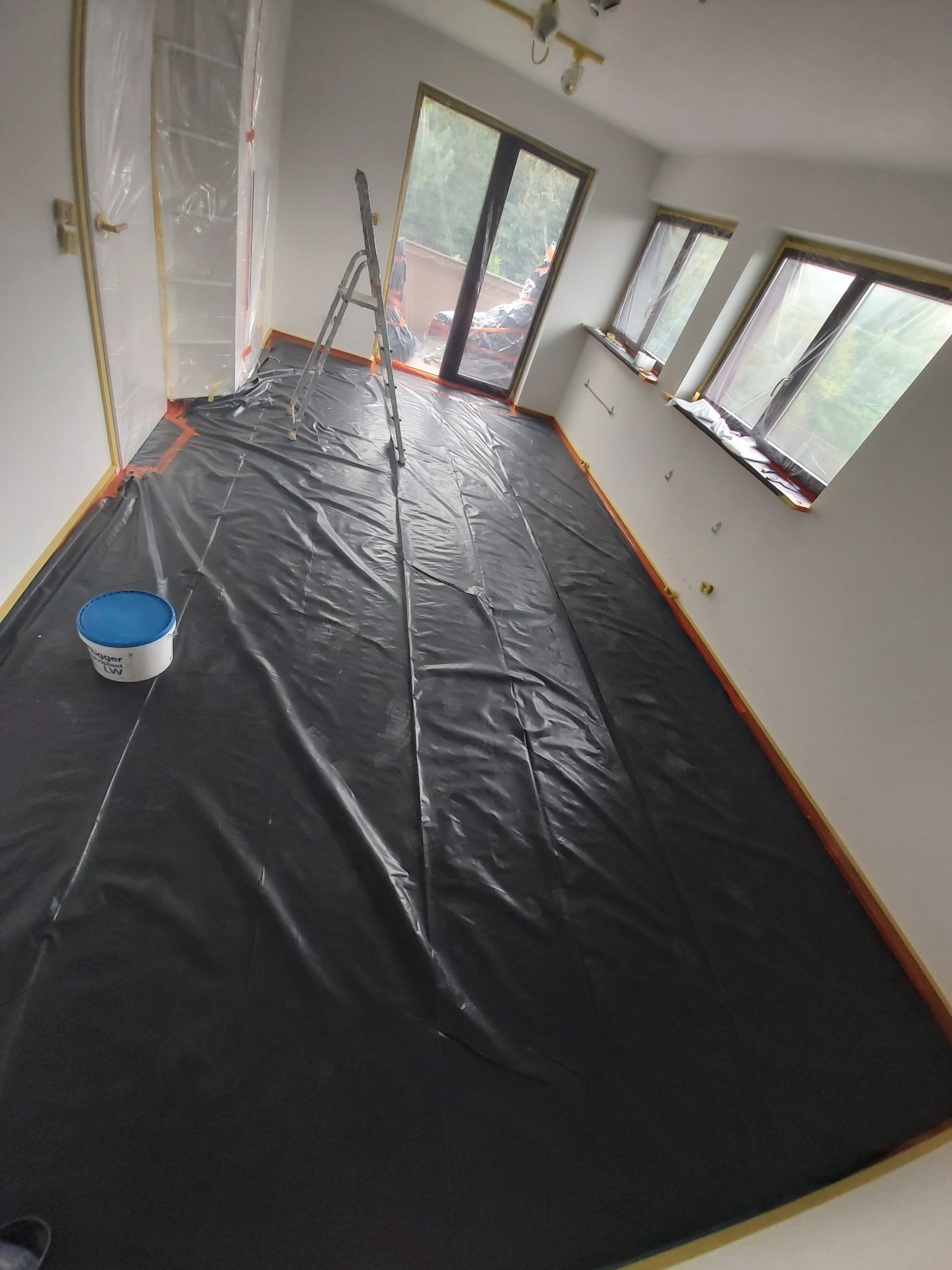 Przygotowanie pokoju do malowania: podłoga zabezpieczona folią, okna i drzwi oklejone taśmą, drabina w rogu, wiadro z farbą. Widok z góry.