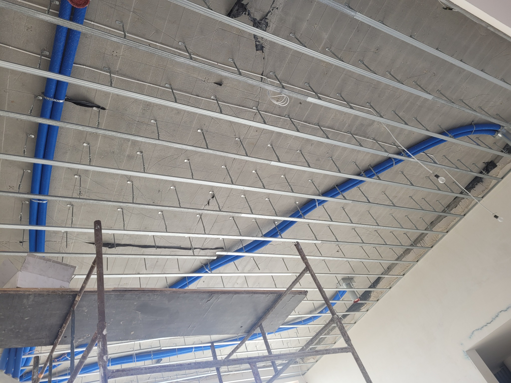 Montaż sufitu podwieszanego z widocznymi elementami konstrukcyjnymi i niebieskimi rurami instalacyjnymi. Widok z dołu, rusztowanie w kadrze.