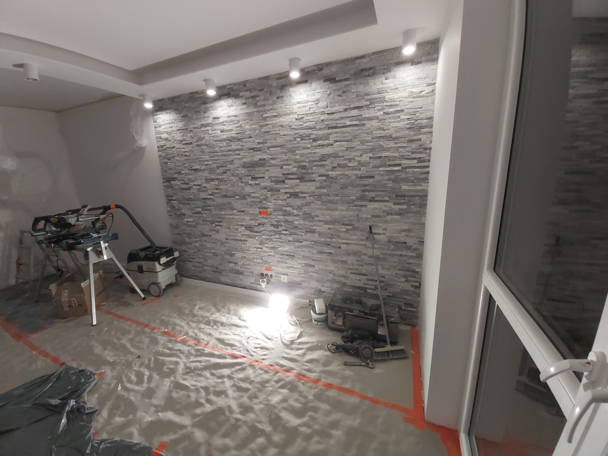 Wykończona ściana z kamienia dekoracyjnego w pokoju, zabezpieczona folią podłoga, narzędzia i sprzęt do cięcia płytek widoczne w tle. Wnętrze w trakcie prac wykończeniowych.