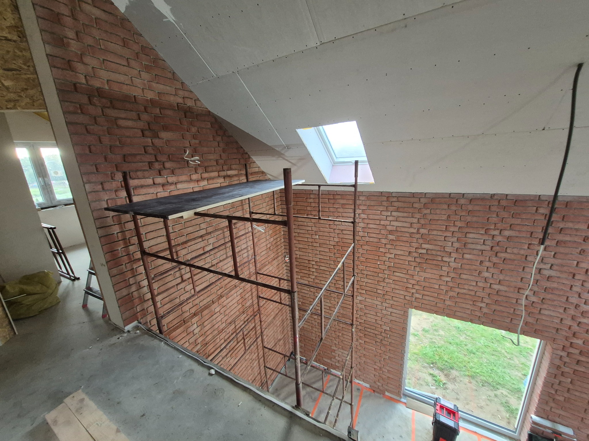 Montaż cegieł dekoracyjnych na ścianie poddasza, widoczne rusztowanie i okno dachowe. Częściowo wykończona ściana z płyt kartonowo-gipsowych.