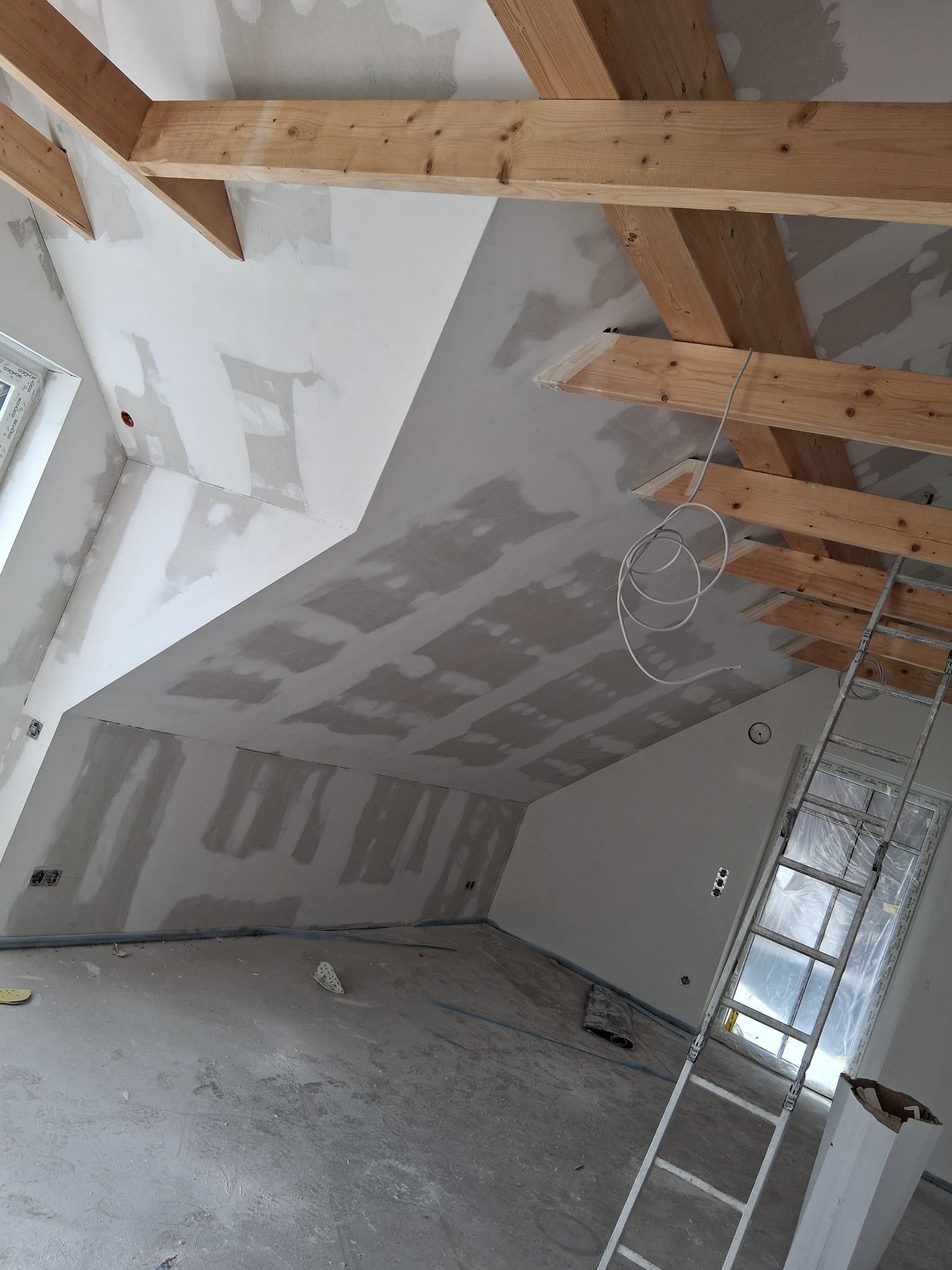 Poddasze w trakcie szpachlowania, widoczne drewniane belki stropowe, ściany pokryte szpachlą, drabina oparta o okno zabezpieczone folią. Wnętrze w stanie surowym.