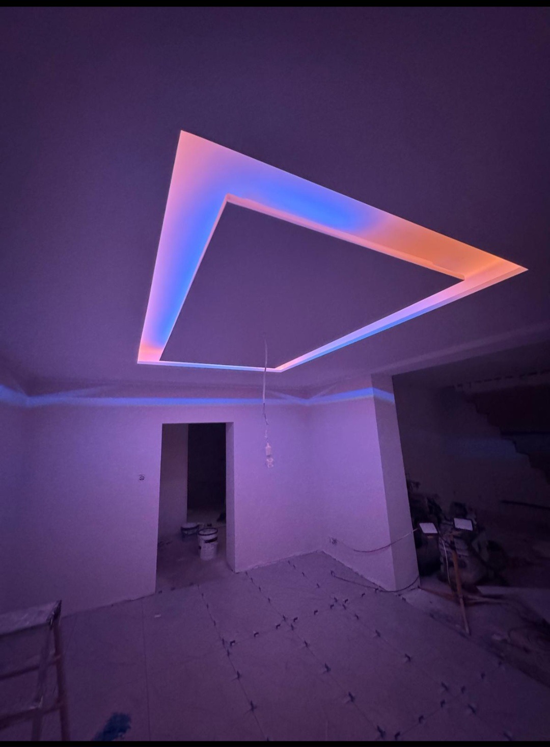 Nowoczesny pokój z podwieszanym sufitem w kształcie kwadratu, oświetlonym niebiesko-pomarańczowym światłem. Na podłodze płytki, ściany białe, w tle widoczne schody.