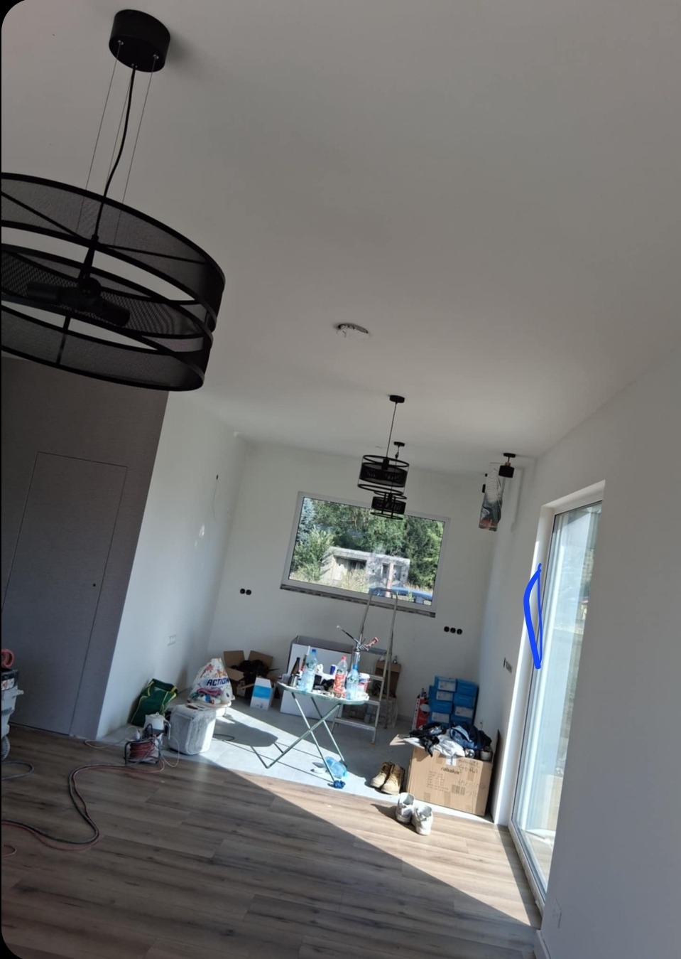 Wnętrze w trakcie prac wykończeniowych: białe ściany, drewnopodobna podłoga, nowoczesne lampy wiszące, okno z widokiem na zieleń, widoczne narzędzia i materiały budowlane.