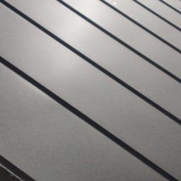 Modern House Mateusz Kuras - Szare panele dachowe z widoczną strukturą, ułożone pod kątem. W tle fragment budynku i zieleń. Widoczne drewniane elementy konstrukcji dachu.