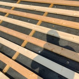 Modern House Mateusz Kuras - Konstrukcja drewniana dachu z deskami i papą, widok z góry. W tle fragment krajobrazu. Cień osoby na konstrukcji. Detal montażu.