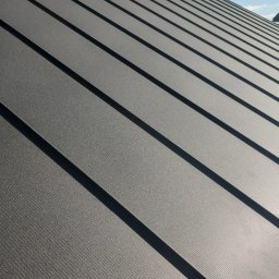 Modern House Mateusz Kuras - Szary dach z blachy na rąbek stojący, z widocznym kominkiem wentylacyjnym i fragmentem niebieskiego nieba. Układ paneli tworzy regularny wzór z wyraźnymi cieniami.