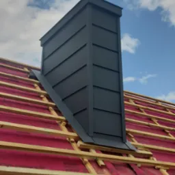 Nowoczesny, grafitowy komin na dachu w trakcie budowy, z widoczną membraną dachową i drewnianą konstrukcją więźby. Kąt ujęcia od dołu podkreśla geometrię komina.