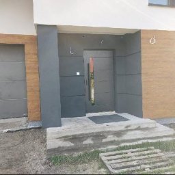 Modern House Mateusz Kuras - Nowoczesny dom z szarą, minimalistyczną fasadą i drewnopodobną okładziną. Wejście z betonowymi schodami i szarą bramą garażową.