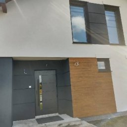 Modern House Mateusz Kuras - Nowoczesny dom z białą elewacją, antracytowymi oknami i drzwiami. Widoczny fragment drewnianej okładziny elewacyjnej. Ujęcie z dołu, podkreślające bryłę budynku.