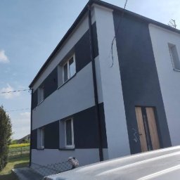 Modern House Mateusz Kuras - Dwukondygnacyjny dom z elewacją w odcieniach szarości, z ciemnymi pasami i jasnymi ścianami. Widoczne okna, drzwi wejściowe oraz fragment dachu. W tle zieleń i błękitne niebo.