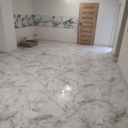 Modern House Mateusz Kuras - Wykończone wnętrze z marmurową podłogą i płytkami na ścianie. Widoczna drewniana stolarka drzwiowa i sufitowe oświetlenie. W tle kartonowe pudło.