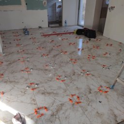 Modern House Mateusz Kuras - Układanie płytek podłogowych z użyciem systemu poziomowania. Widoczne narzędzia i materiały budowlane. Jasne płytki z marmurkowym wzorem.