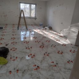 Modern House Mateusz Kuras - Wnętrze w trakcie remontu: ułożone marmurowe płytki podłogowe z pomarańczowymi klipsami poziomującymi, drabina oparta o okno, ściany w stanie surowym, widoczne narzędzia.