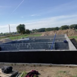 Modern House Mateusz Kuras - Wykopane fundamenty pod dom z izolacją przeciwwilgociową i częściowo wymurowanymi ścianami z bloczków betonowych na tle krajobrazu polnych wzgórz.