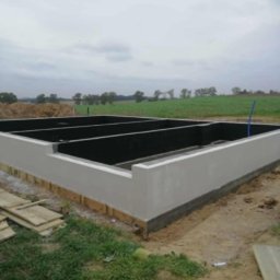 Modern House Mateusz Kuras - Zalane fundamenty domu z widoczną hydroizolacją wewnątrz, na zewnątrz drewniane deski i ziemia z trawą. W tle zielone pole i pochmurne niebo.