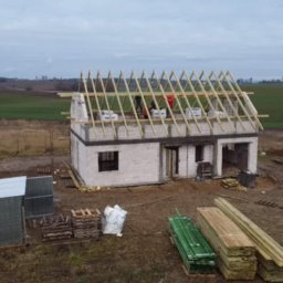 Modern House Mateusz Kuras - Budowa domu jednorodzinnego w stanie surowym otwartym: ściany z betonu komórkowego, drewniana konstrukcja dachu, materiały budowlane na działce. Widok z lotu ptaka.