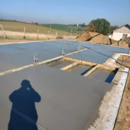 Świeżo wylana, gładka płyta fundamentowa z wystającymi prętami zbrojeniowymi, otoczona wykopem. W tle pagórkowaty krajobraz i częściowo widoczny budynek.