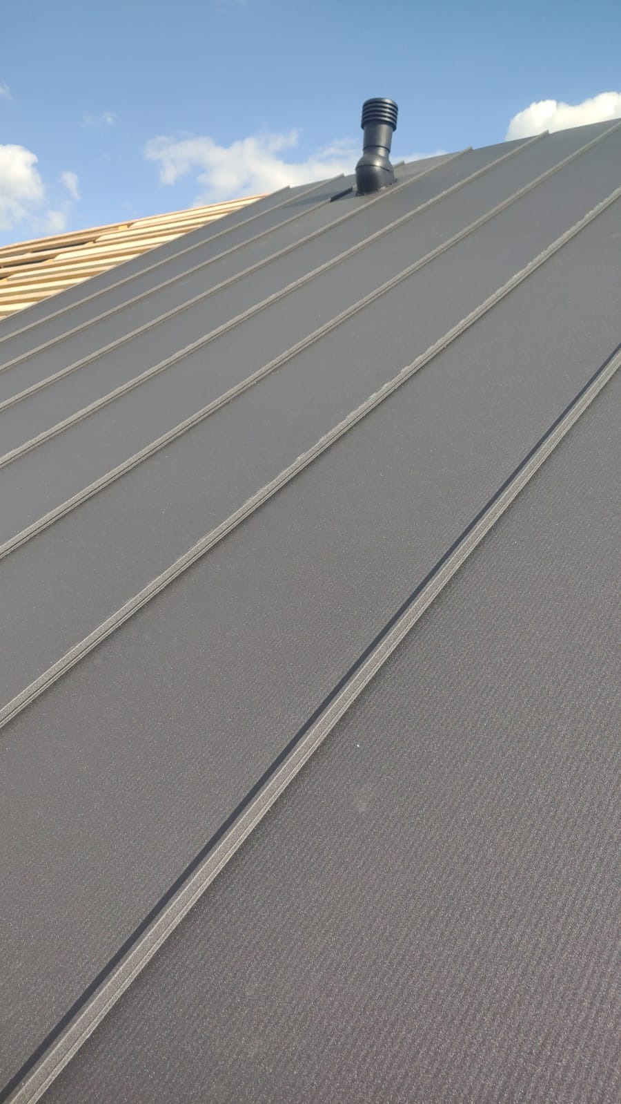 Szary dach z blachy na rąbek stojący, z kominkiem wentylacyjnym i widoczną konstrukcją drewnianą. Ujęcie z dołu, błękitne niebo w tle.