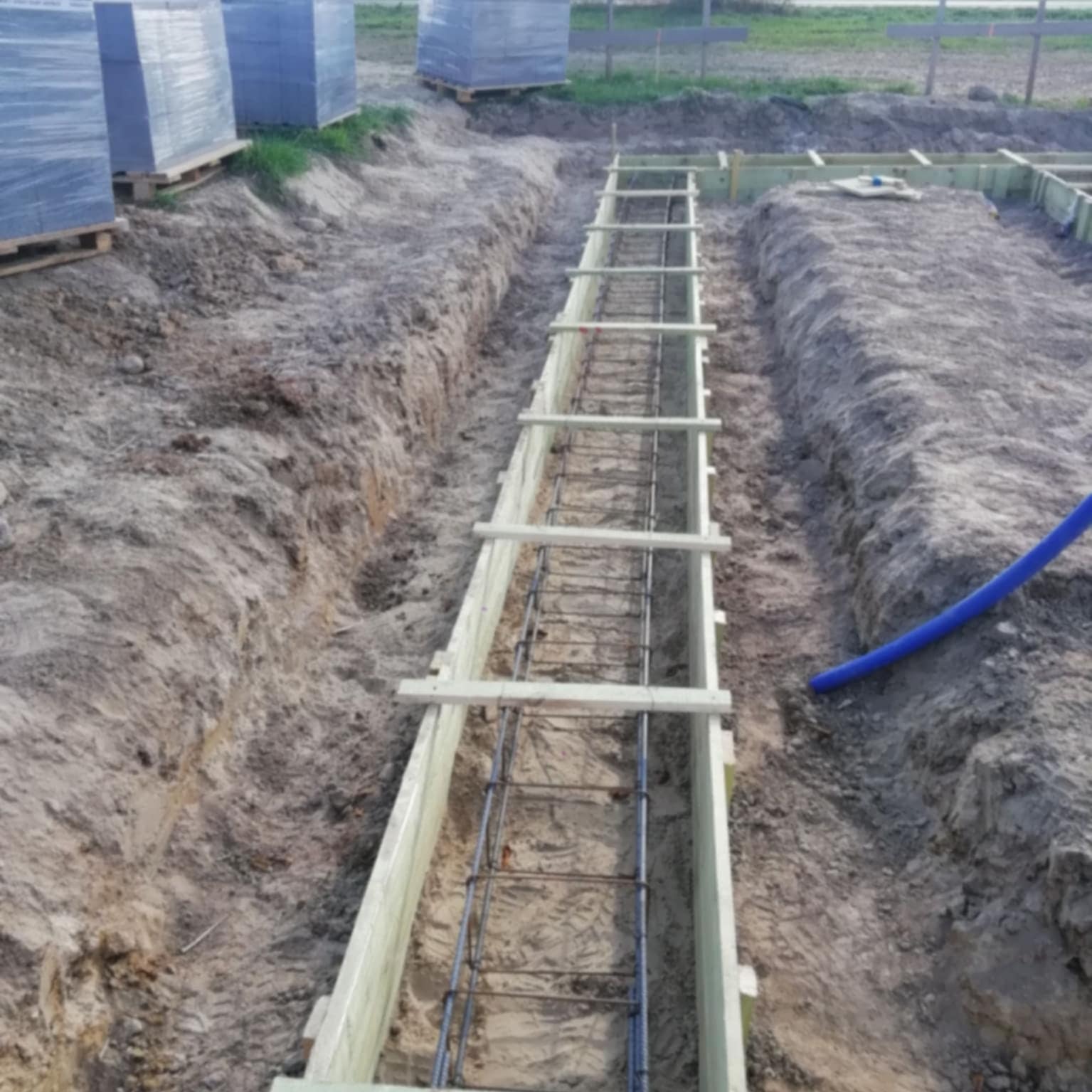 Przygotowanie wykopu pod fundamenty z szalunkiem i zbrojeniem. W tle palety z bloczkami betonowymi. Widok z góry na plac budowy domu.